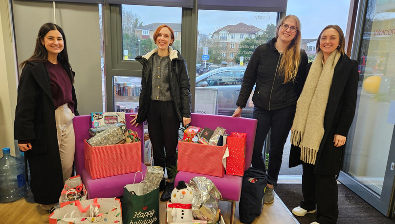 The picture shows four women stood up and surrounded by gift-wrapped Christmas presents