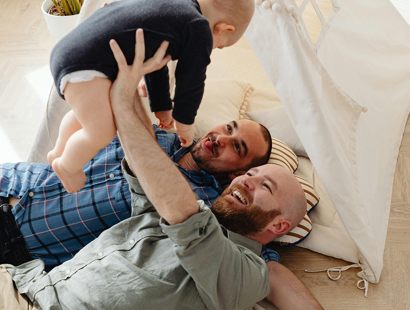 Two men holding up a baby in the air, lying on an indoor tent. One man in a blue chequed shirt; the other in a green linen shirt. Take PRIDE in your estate planning: three considerations for the LGBTQIA+ community