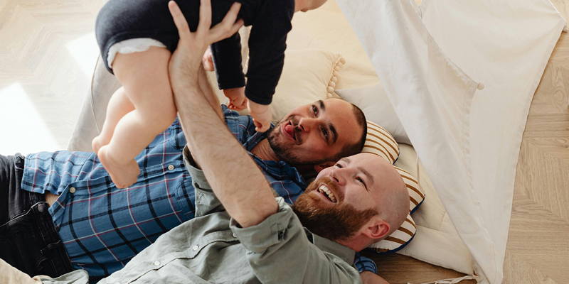 Two men holding up a baby in the air, lying on an indoor tent. One man in a blue chequed shirt; the other in a green linen shirt. Take PRIDE in your estate planning: three considerations for the LGBTQIA+ community