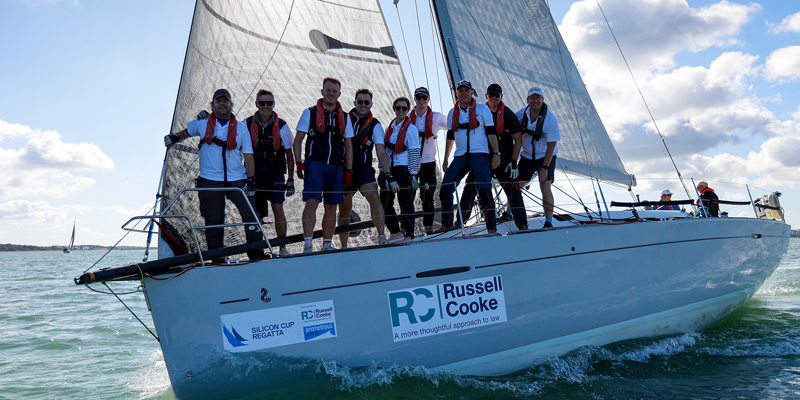 The picture shows a sailing boat with nine people standing aboard. The boat has the Russell-Cooke logo on the side of the boat. 