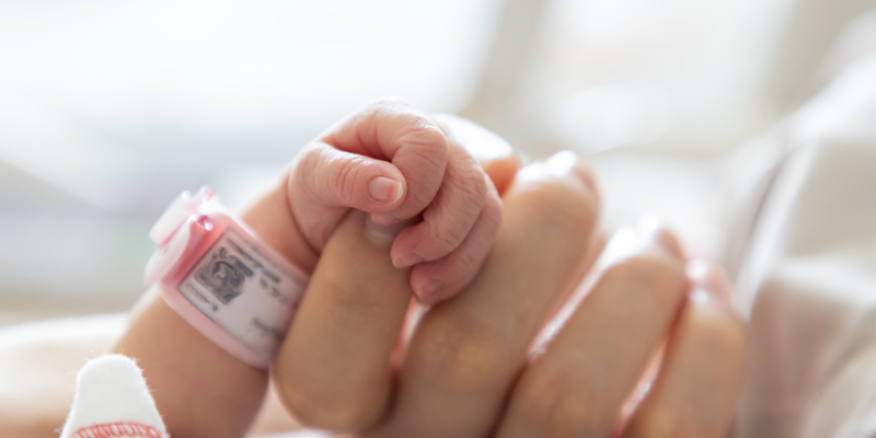 A newborn baby's arm and hand with identification band on its wrist, holding an adult hand. Continuing the conversation around birth trauma and childbirth-related PTSD