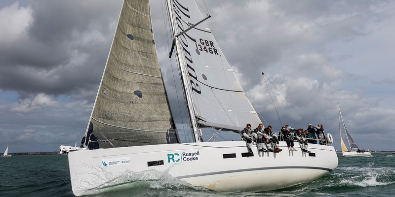 A white sailing boat afloat in the sea with five individuals sitting on the edge. Russell-Cooke crew aboard a sailing boat at the Silicon Cup Regatta 2025