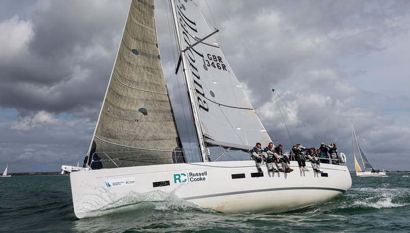 A white sailing boat afloat in the sea with five individuals sitting on the edge. Russell-Cooke crew aboard a sailing boat at the Silicon Cup Regatta 2025