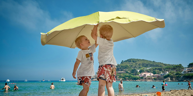 Two young boys putting up a parasol on a beach with the Mediterranean sea behind them. Summer Holidays: who has what time with the children?