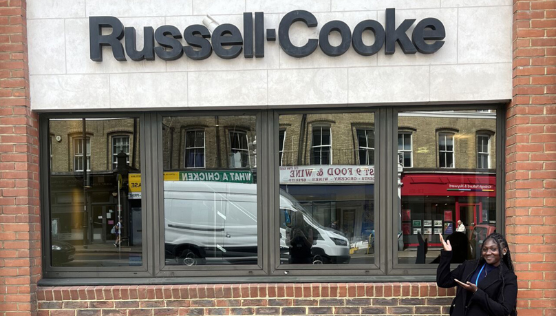 A red-bricked building with with brown windows and a black signage spelling 'Russell-Cooke' with a black female on the far right pointing to the Russell-Cooke sign. Aspiring solicitor Moriah Joseph-Brown in front of the Russell-Cooke sign outside the Putney office.