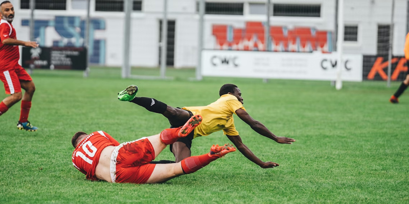 The image shows two football players on the floor having fallen over in a tackle. There is a football player running in the background
