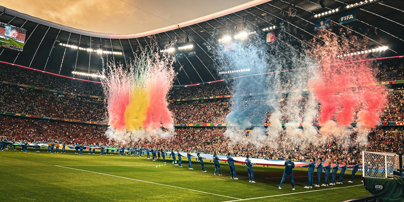 The picture shows pre-match celebrations ahead of a World Cup Game. There are flares going off and players holding a flag on the pitch in the stadium. 